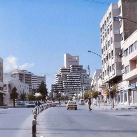 شارع في منطقة الشميساني بعمّان في ثمانينات القرن الماضي، تظهر فيه مبانٍ مكتبية ومبنى مدرّج مرتفع في الخلفية